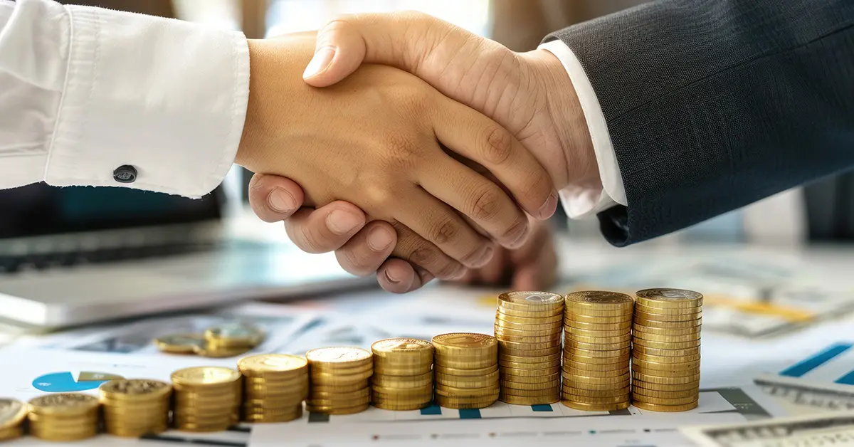 Businesspeople shaking hands over increasing stacks of coins and financial documents