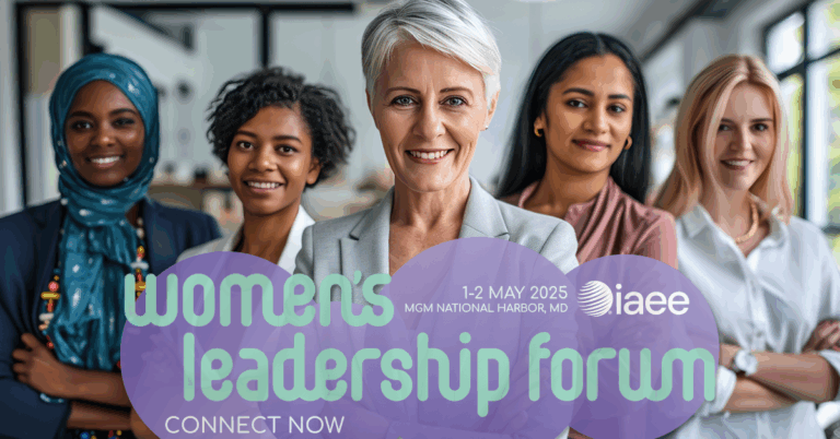 Group of diverse professional women confidently standing in a line in front of 2025 IAEE Women's Leadership Forum logo, with the woman in the foreground crossing her arms, showcasing a strong and united front in a workplace setting.